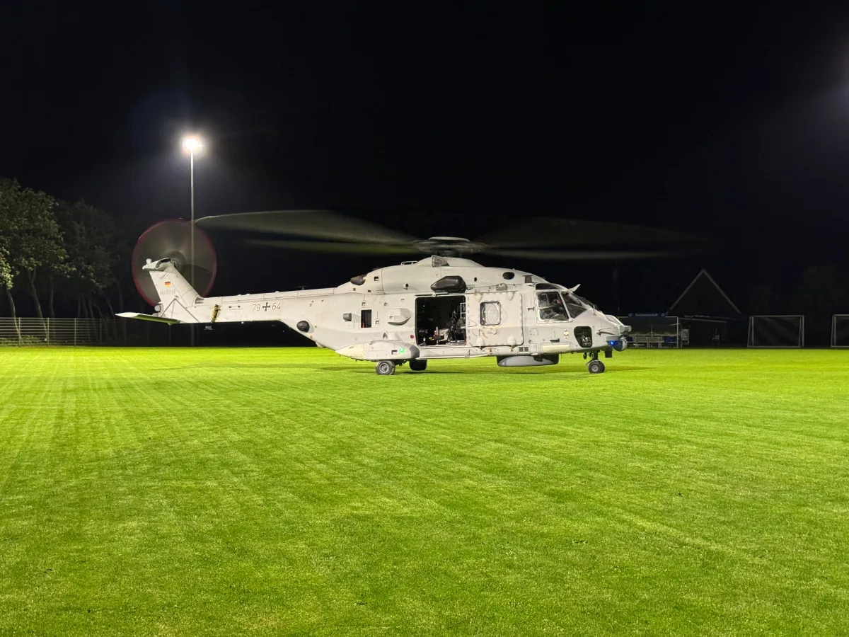 Ein großer, weißer Hubschrauber steht nachts mit rotierenden Rotorblättern auf einem beleuchteten Sportplatz. Bäume und ein Haus sind im Hintergrund sichtbar.