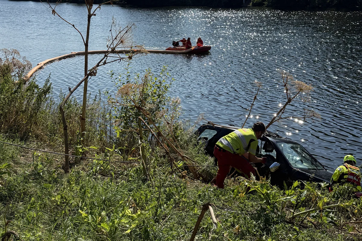 Ein Auto ist teilweise im Wasser versunken; daneben arbeiten zwei Rettungskräfte. Ein Rettungsboot ist auf dem Fluss im Einsatz.