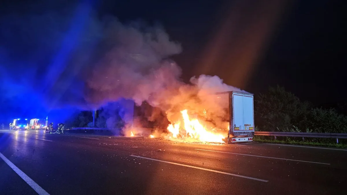 Ein Lkw brennt auf einer dunklen Autobahn, umgeben von Feuerwehrleuten und blauem Licht der Einsatzfahrzeuge.