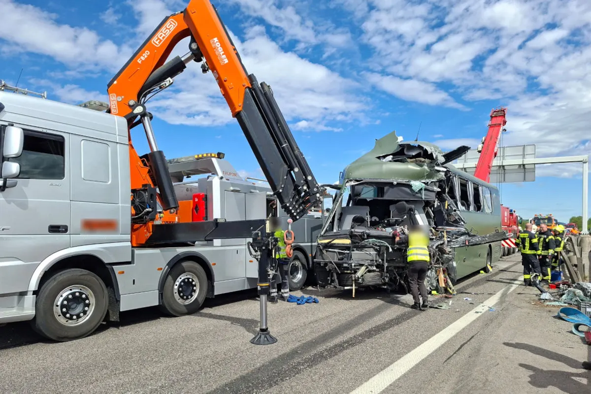 Ein stark beschädigter Bus steht auf einer Autobahn, flankiert von einem silbernen Kranfahrzeug. Mehrere Personen in gelben Westen sind anwesend.