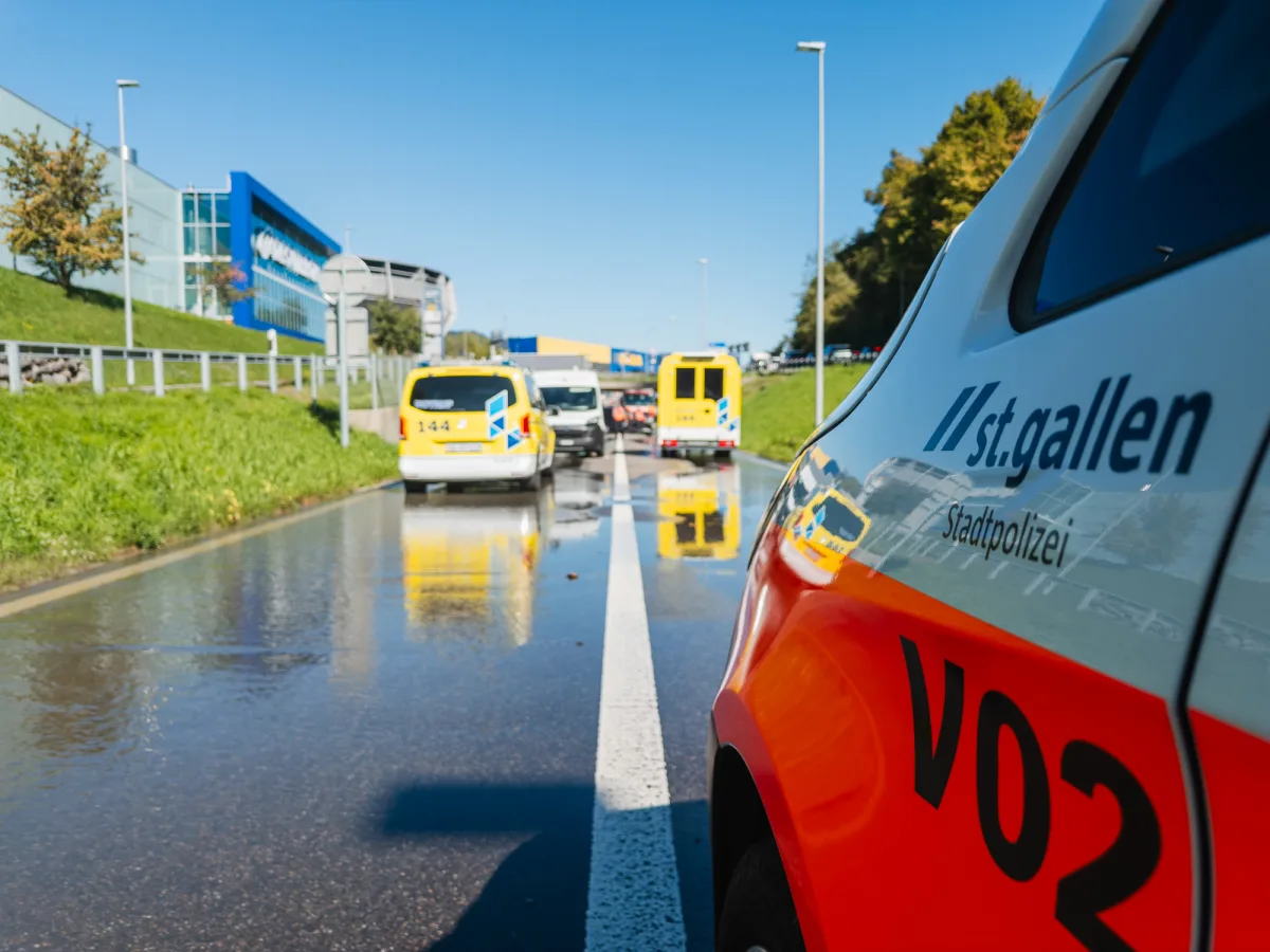 Ein Fahrzeug der Stadtpolizei St. Gallen steht auf einer sonnigen Straße mit großen Pfützen. Im Hintergrund sind weitere Einsatzfahrzeuge zu sehen.