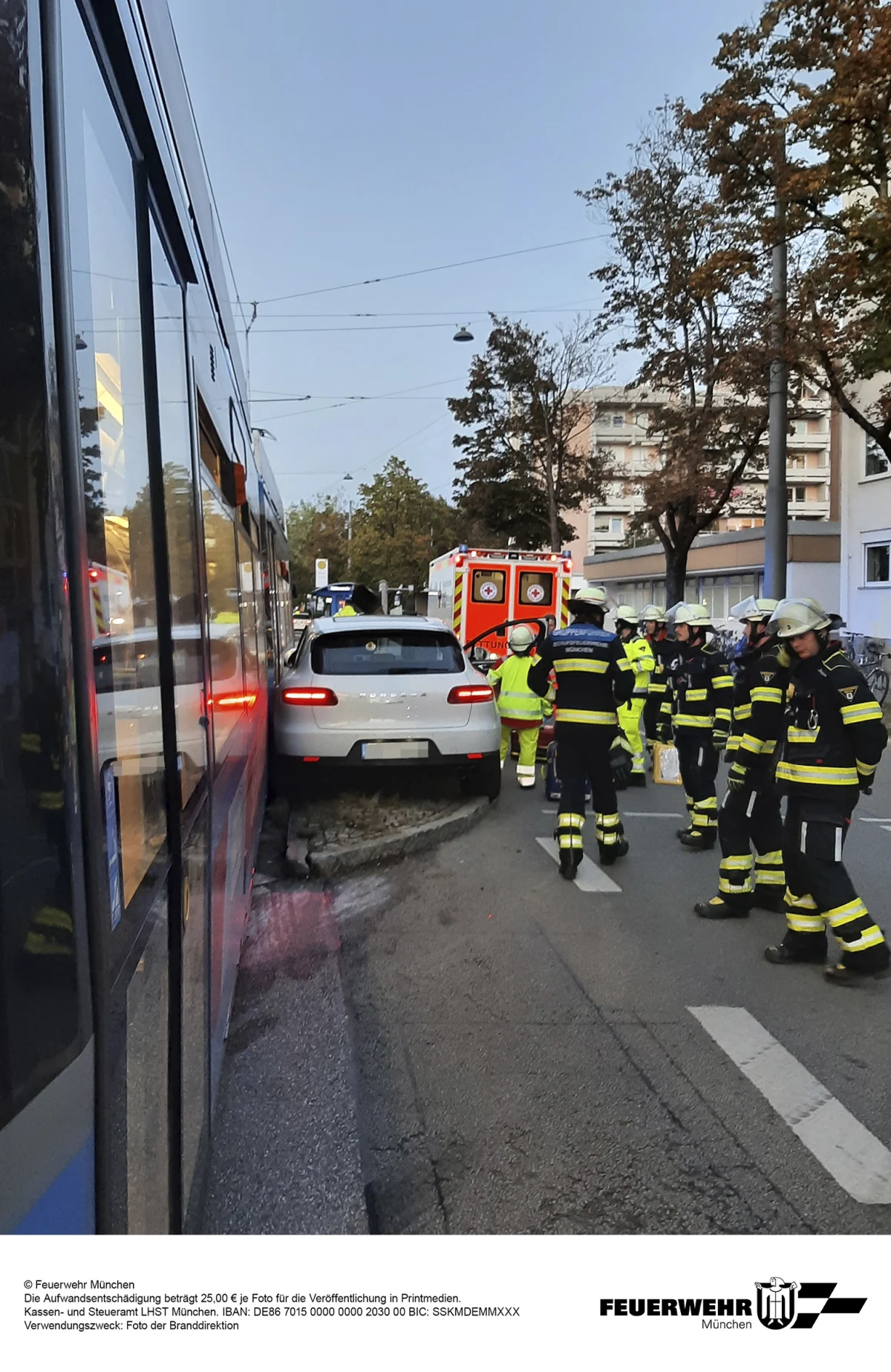 Ein weißes Auto ist zwischen einer Straßenbahn und einem Bordstein eingeklemmt. Feuerwehrleute und ein Rettungswagen sind vor Ort.