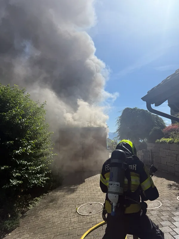 Ein Feuerwehrmann in Schutzkleidung steht vor einem Gebäude, aus dem dichter Rauch aufsteigt. Es ist ein sonniger Tag, und er hält einen Wasserschlauch in der Hand.