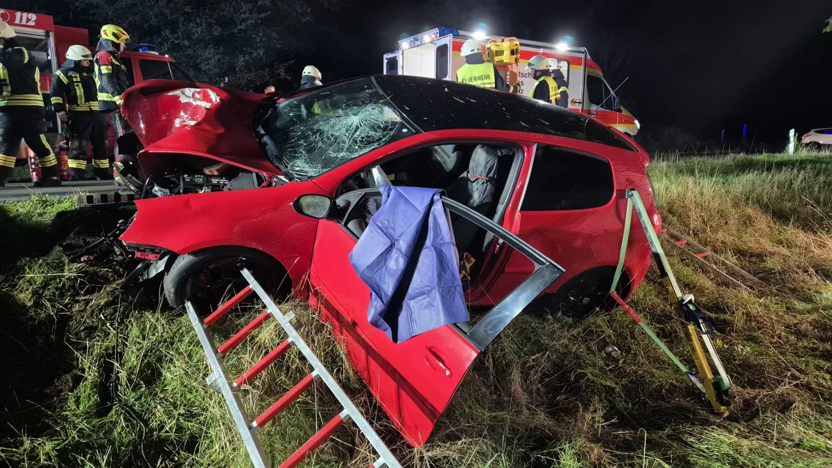 Das Bild zeigt ein stark beschädigtes rotes Auto in einer Graslandschaft bei Nacht. Im Hintergrund sind Rettungskräfte und ein Feuerwehrfahrzeug sichtbar.