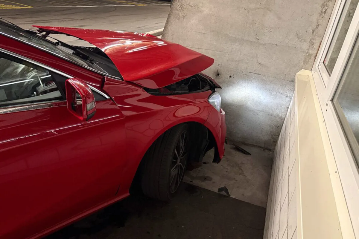 Ein rotes Auto ist frontal mit einem Betonpfeiler kollidiert, wobei die Motorhaube hochgebogen ist. Die Szene befindet sich in einer Parkgarage.