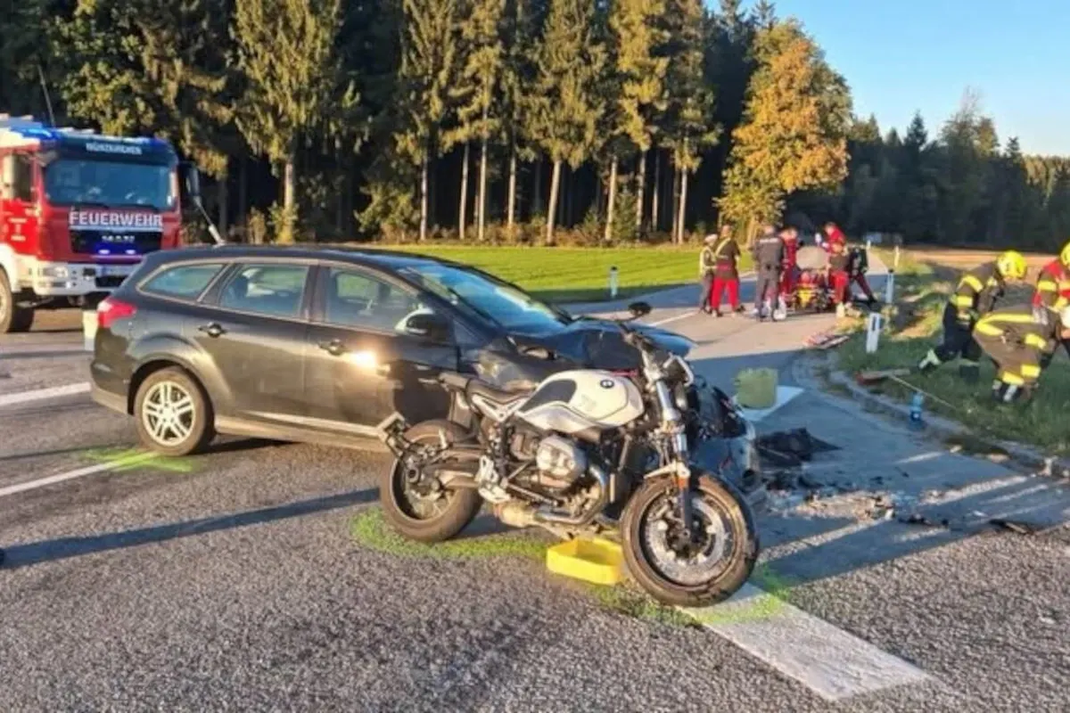 Ein Motorrad steht beschädigt vor einem dunklen Auto an einer Kreuzung. Im Hintergrund ist die Feuerwehr vor Ort.