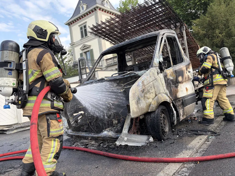 Zwei Feuerwehrmänner in voller Ausrüstung bekämpfen das Feuer in einem ausgebrannten Lieferwagen. Im Hintergrund steht ein großes, historisches Gebäude.