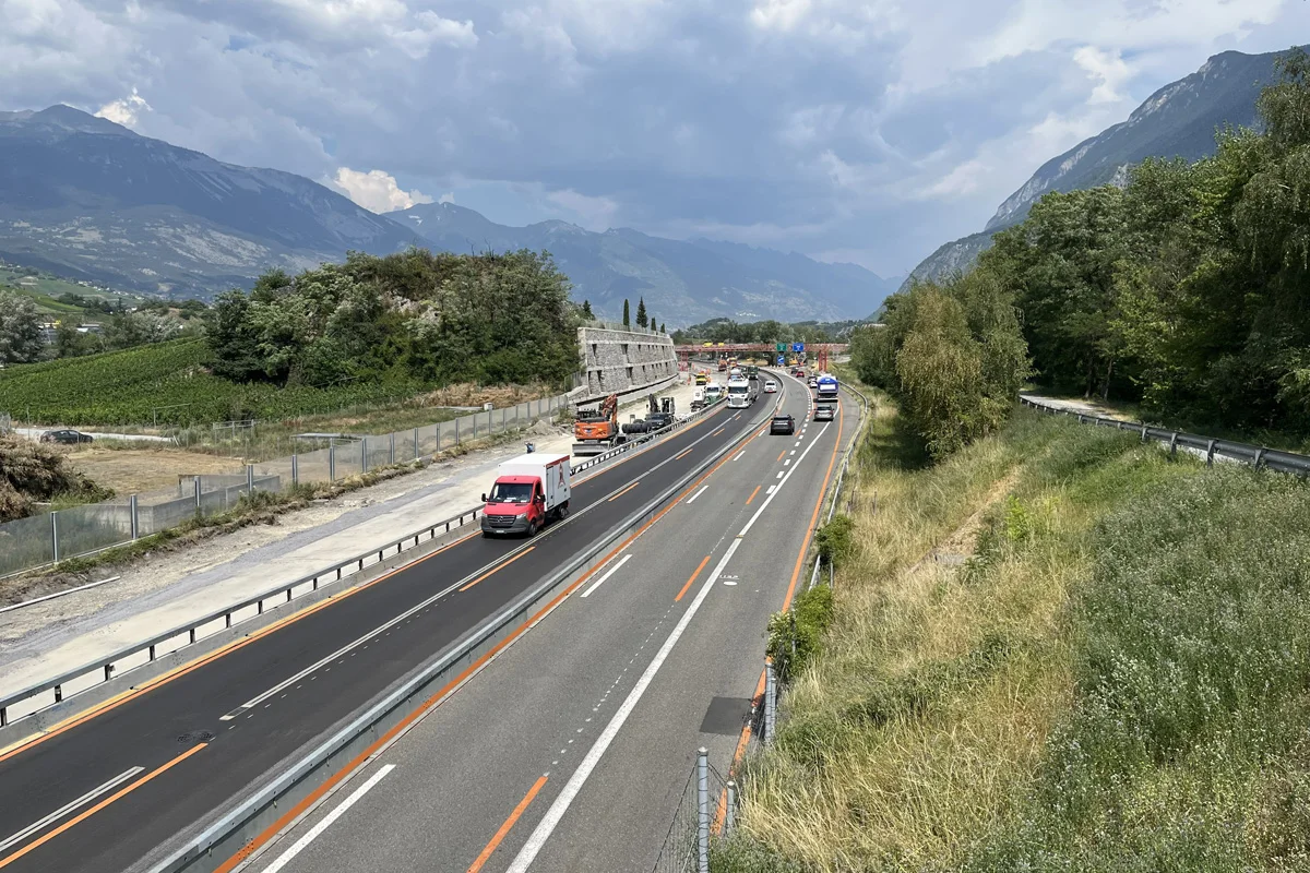 Eine Straße mit mehreren Fahrzeugen, darunter ein roter Lieferwagen, verläuft durch eine bergige, grüne Landschaft. Der Himmel ist mit Wolken bedeckt.