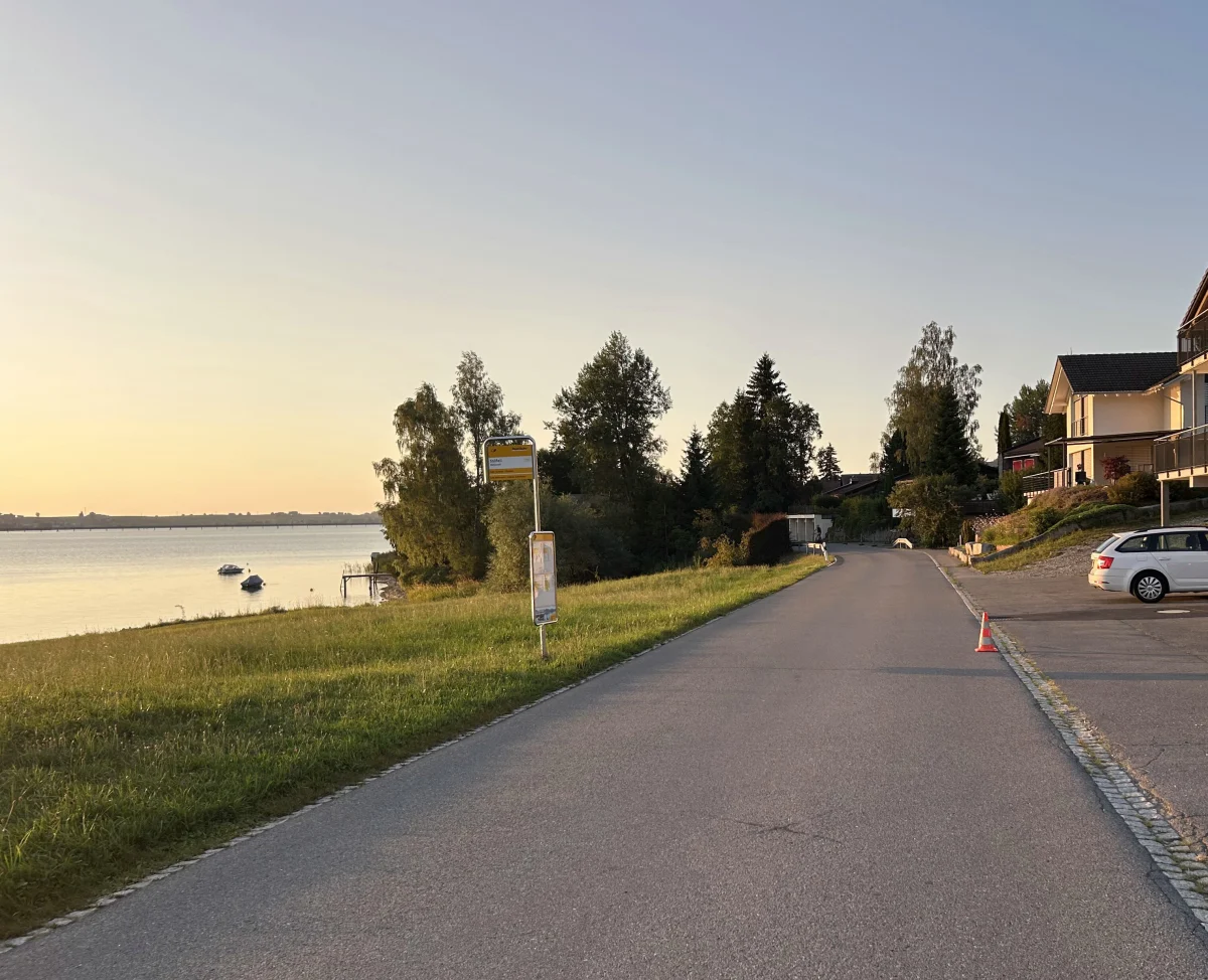Eine ruhige Straße mit einem Verkehrskegel und Häusern, die am Ufer eines Sees in der Abenddämmerung liegen. Im Hintergrund sind Boote auf dem Wasser zu sehen.