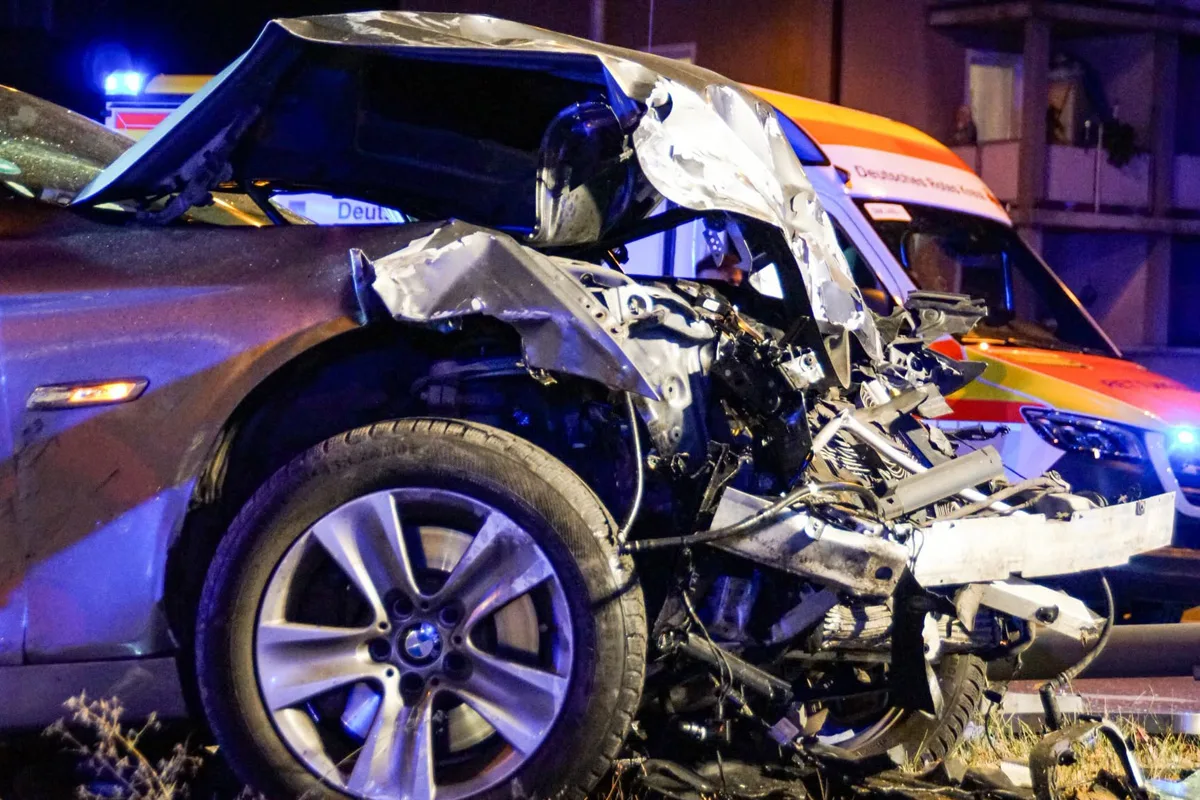 Ein stark beschädigtes Auto nach einem Unfall bei Nacht, im Hintergrund ist ein Rettungsfahrzeug zu sehen. Teile des Wagens sind verformt und zeigen deutliche Unfallspuren.