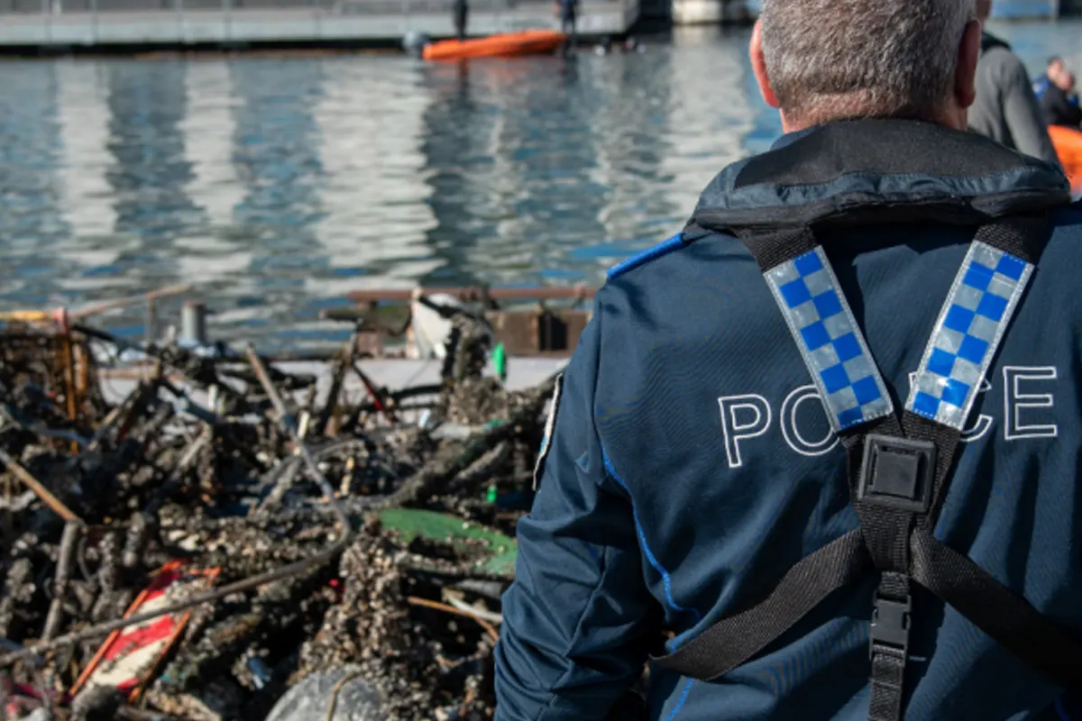Ein Polizist in Uniform steht auf einem Steg und blickt auf eine Ansammlung von geborgenem Müll. Im Hintergrund ist ein Gewässer zu sehen.