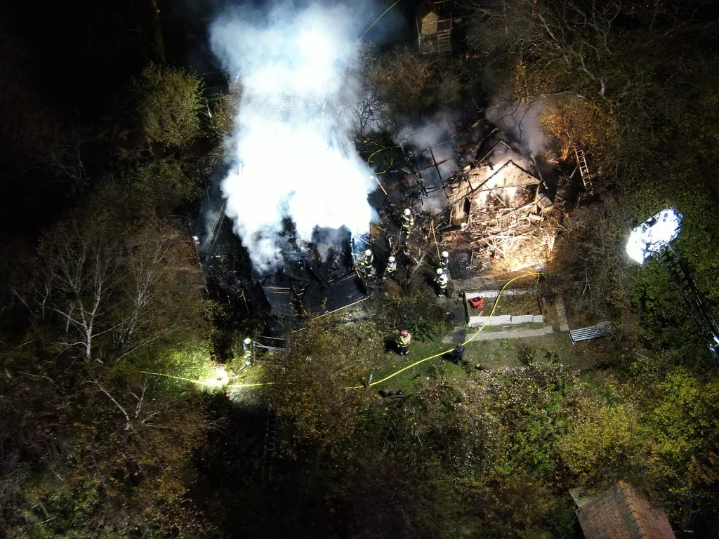 Das Bild zeigt eine Luftaufnahme von Feuerwehrleuten, die einen Brand bekämpfen. Ein Gebäude in einer Waldlichtung ist zerstört und raucht stark.