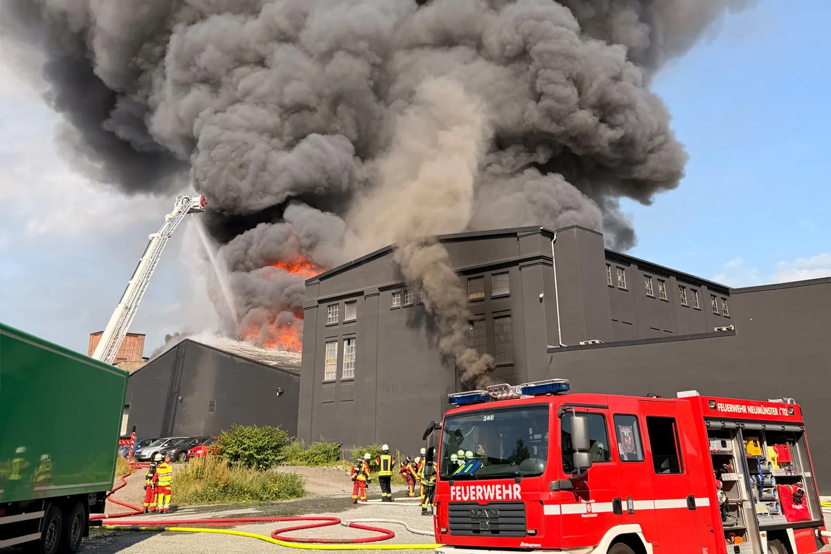 Ein Feuerwehrfahrzeug und Einsatzkräfte sind bei einer brennenden Lagerhalle zu sehen. Dichter Rauch steigt auf.