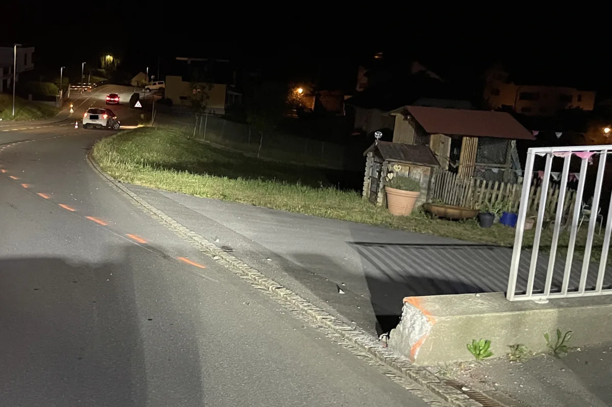Zwei Autos auf einer nächtlichen Straße; eines parkt nahe einem Betonpfeiler mit sichtbaren Schäden. Straßenbeleuchtung und Häuser im Hintergrund.
