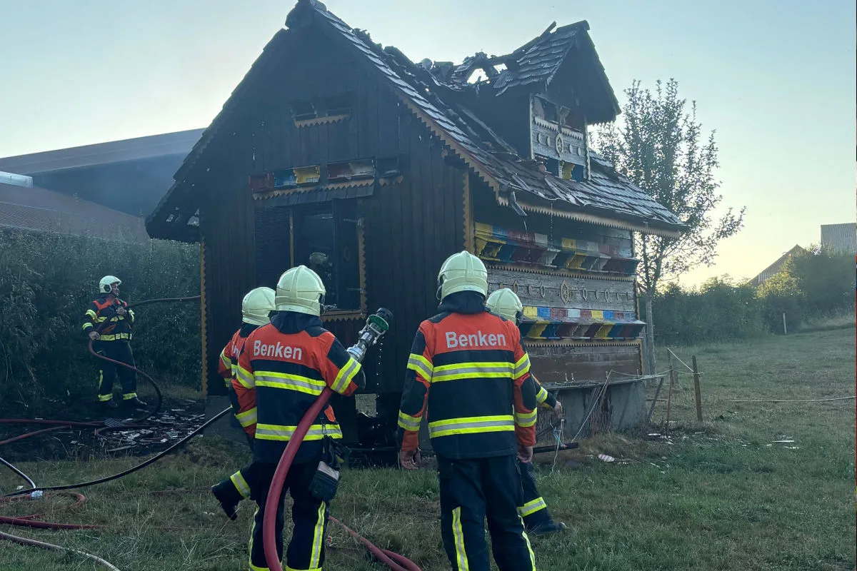 Mehrere Feuerwehrleute in Uniformen mit 'Benken'-Aufschrift löschen ein brennendes Holzgebäude. Der Dachstuhl ist beschädigt.