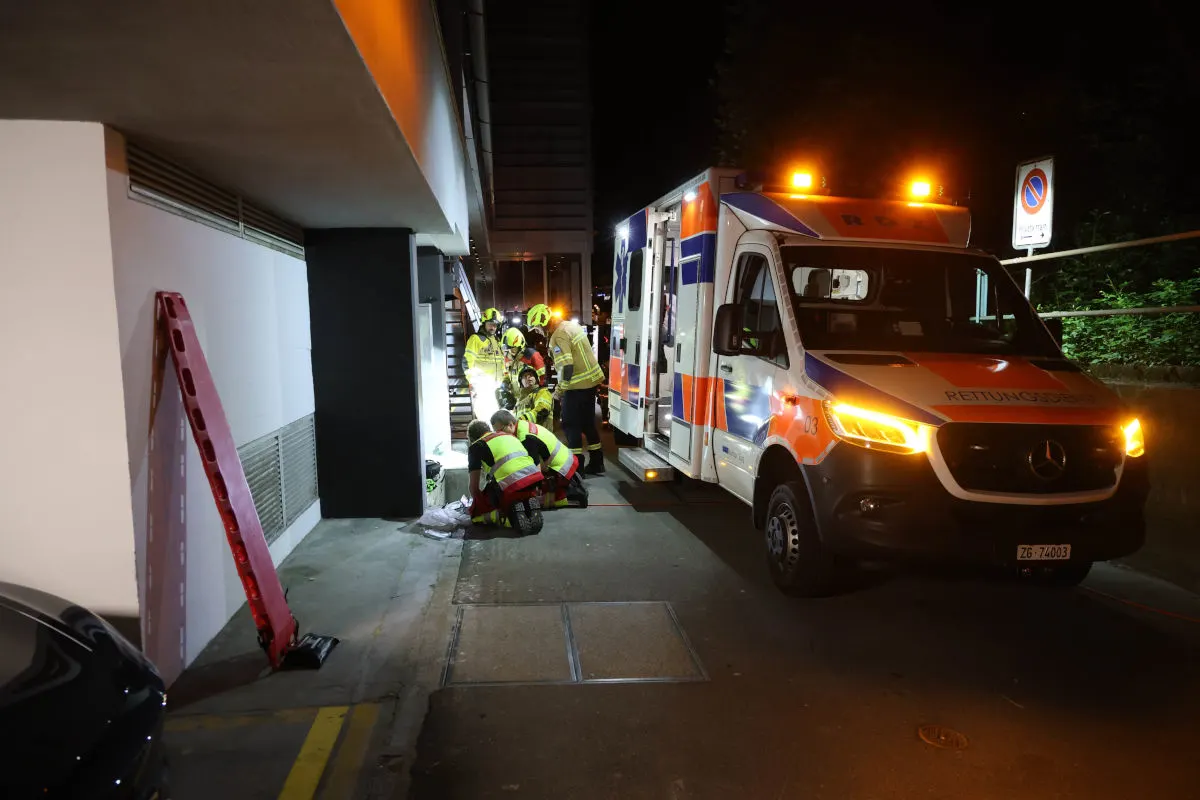Mehrere Einsatzkräfte sind im Einsatz neben einem Krankenwagen mit leuchtenden Frontlichtern. Die Szene ist nachts in einer städtischen Umgebung.