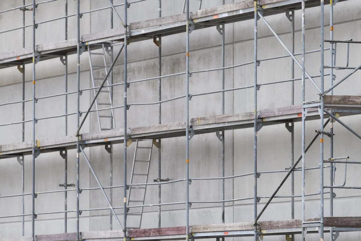 Ein mehrstöckiges Baugerüst ohne Arbeiter vor einer grauen Betonwand. Das Gerüst ist leer.