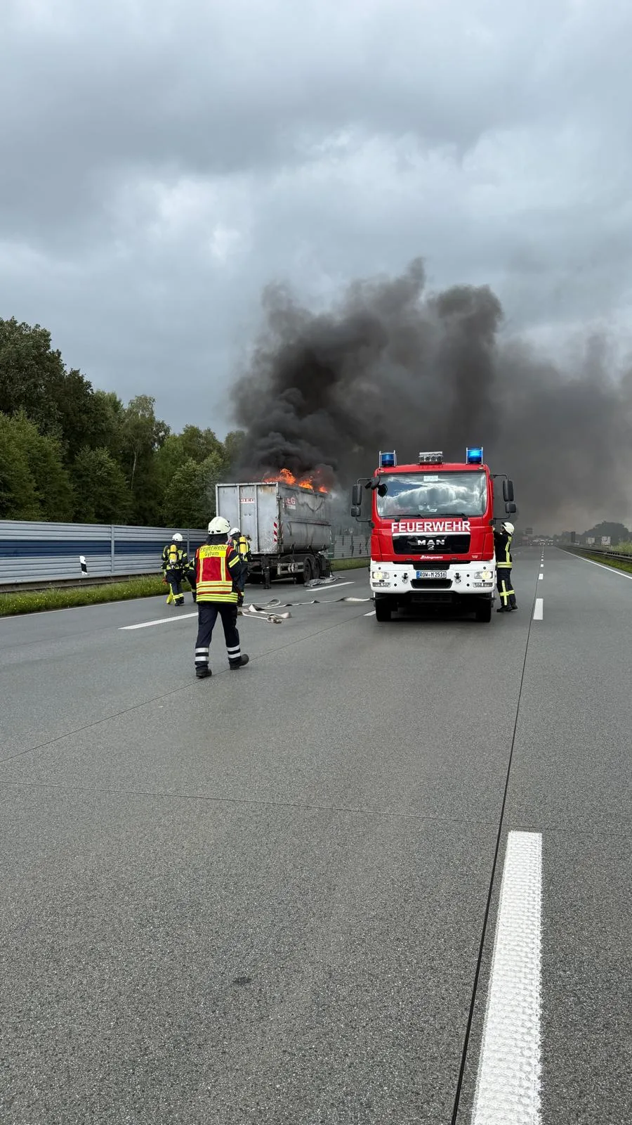 Ein brennender Lastwagen steht auf der Autobahn, umgeben von Rauch. Feuerwehrleute arbeiten an der Löschung.