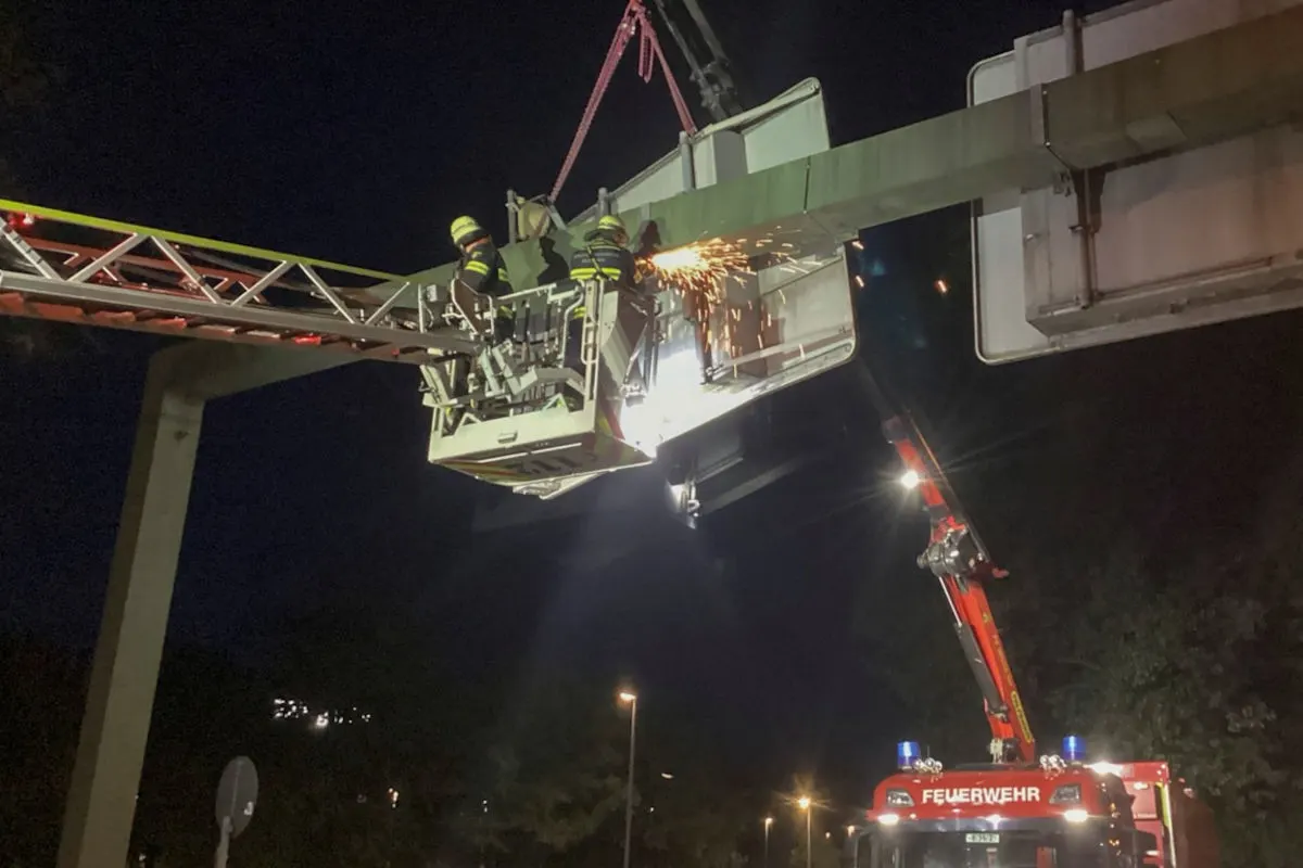 Bei Nacht arbeiten Feuerwehrleute in einem Hebekorb an einem großen Verkehrsschild, mit Funkenflug sichtbar.