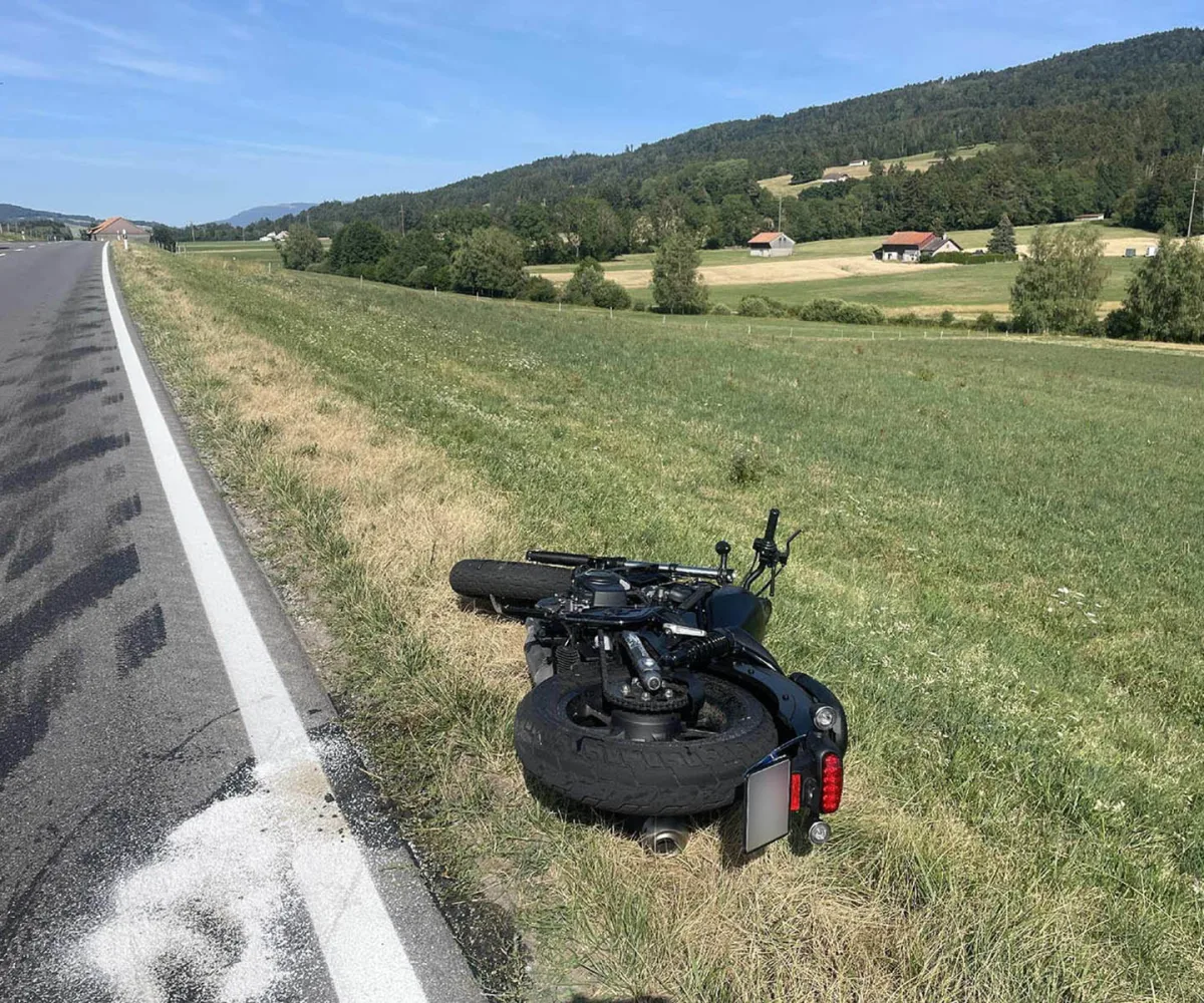 Ein schwarzes Motorrad liegt umgestürzt auf einer Wiese neben einer Landstraße. Die Straße ist geteilt und verläuft durch eine ländliche Gegend mit Feldern und Hügeln im Hintergrund.