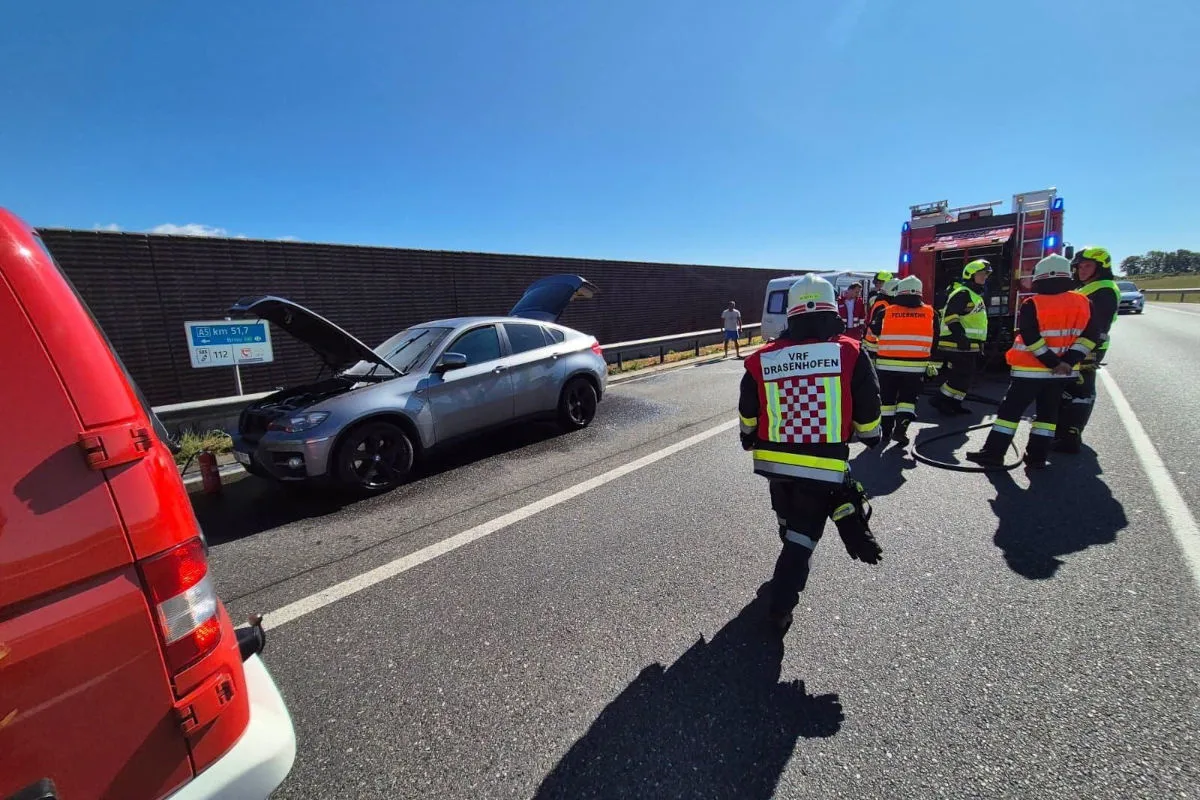 Feuerwehrleute in Einsatzkleidung stehen auf einer Autobahn vor einem Auto mit offener Motorhaube. Ein Einsatzfahrzeug ist zu sehen.