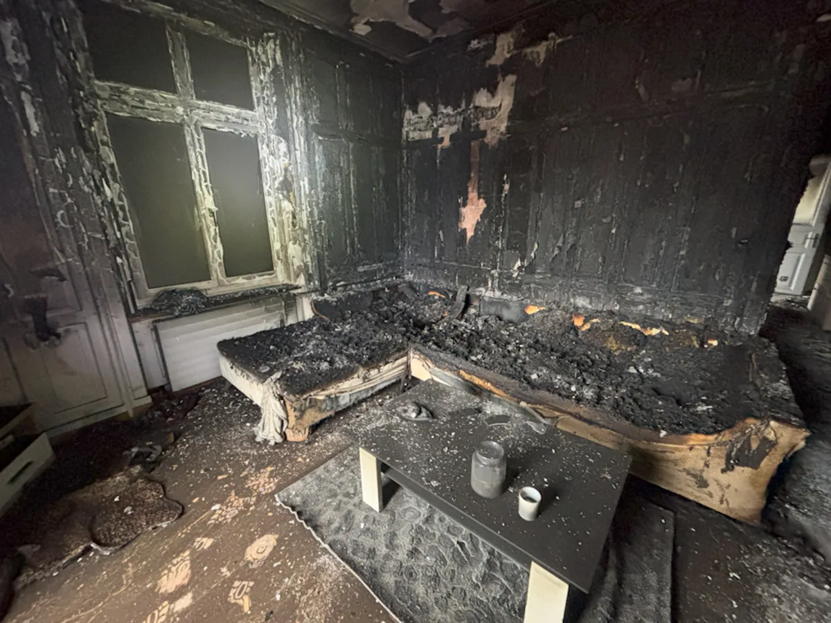 Das Bild zeigt ein komplett ausgebranntes Zimmer mit einem stark zerstörten Sofa vor der schwarzen Wand. Fensterrahmen und Wände sind ebenfalls stark beschädigt.