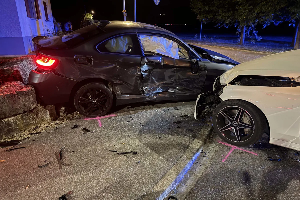 Zwei Fahrzeuge, eines schwarz und das andere weiß, sind nach einer Kollision auf einer Straße beschädigt. Der schwarze Wagen steht seitlich, während der weiße Wagen frontal beschädigt ist.