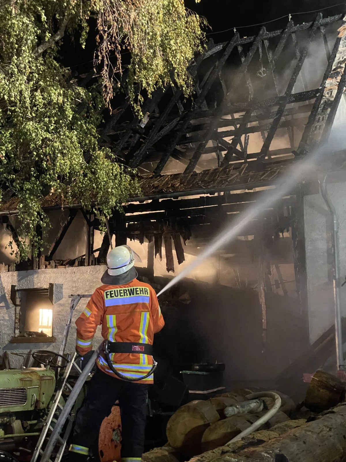 Ein Feuerwehrmann in oranger Uniform sprüht Wasser auf ein von dunklen Rauch umgebenes, brennendes Hausdach.