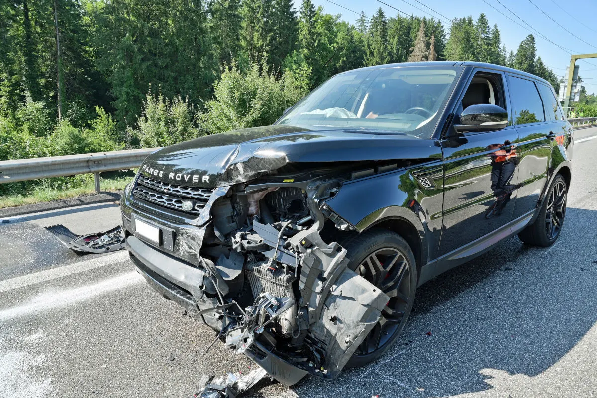 Ein schwarzer Range Rover steht mit schwer beschädigter Front auf einer Landstraße. Im Hintergrund sind grüne Bäume und eine Leitplanke sichtbar.