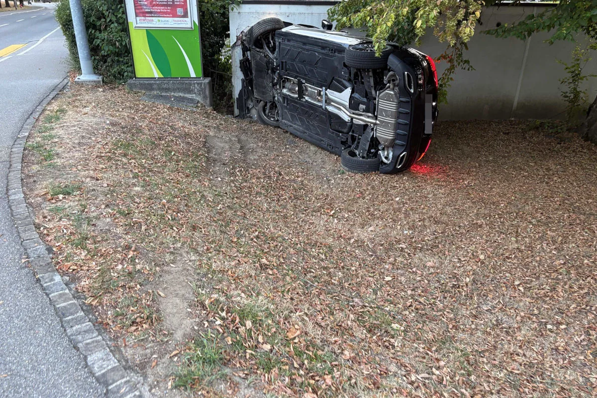 Ein schwarzes Auto liegt auf der Seite neben einer Straße, umgeben von trockenem Gras. Es ist keine aktive Rettung zu sehen.