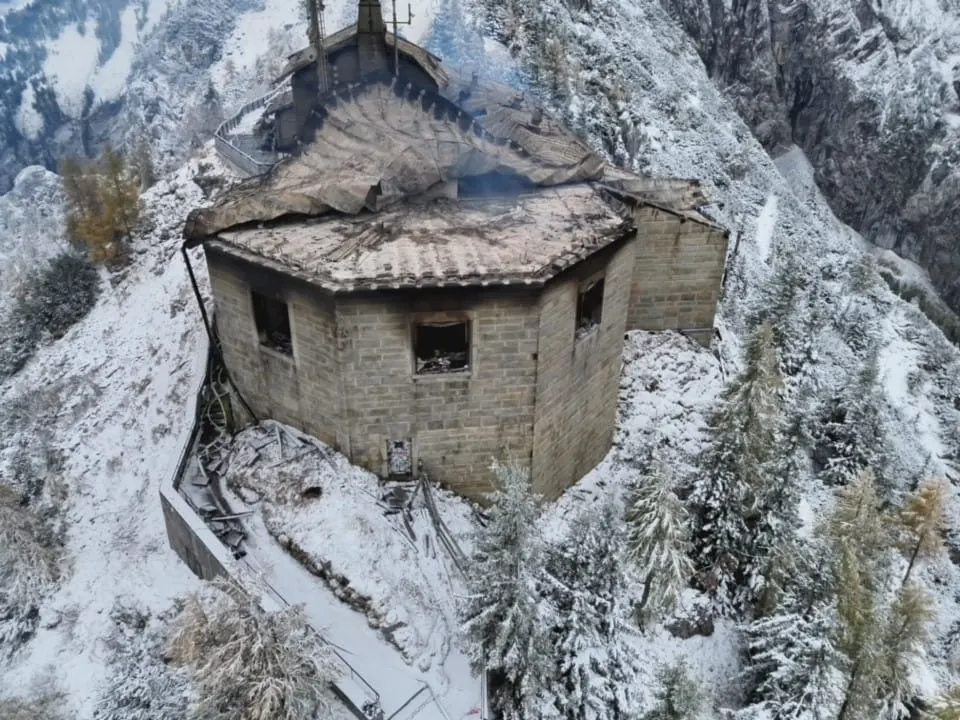 Ein ausgebranntes Gebäude steht auf einem verschneiten Berghang. Das Dach ist teilweise eingestürzt, und Rauchspuren sind sichtbar.