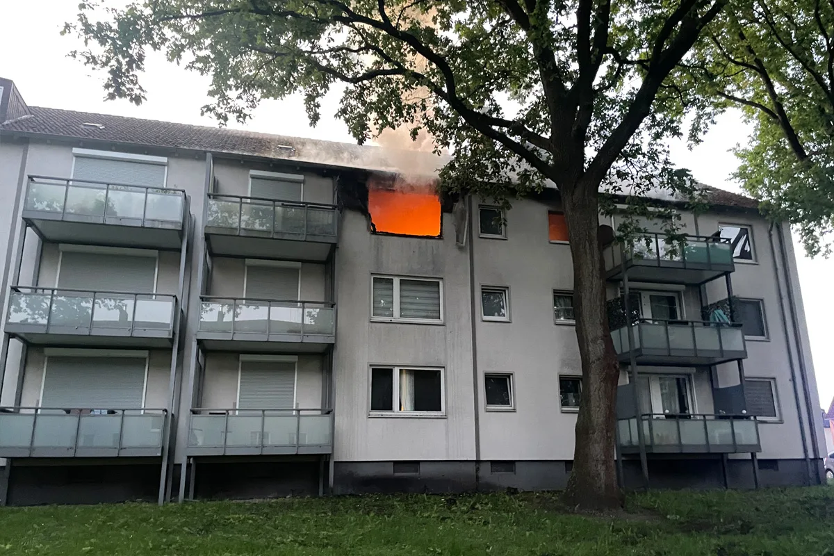 Ein Mehrfamilienhaus mit brennendem Fenster im obersten Stockwerk. Rauch steigt aus dem Gebäude auf.