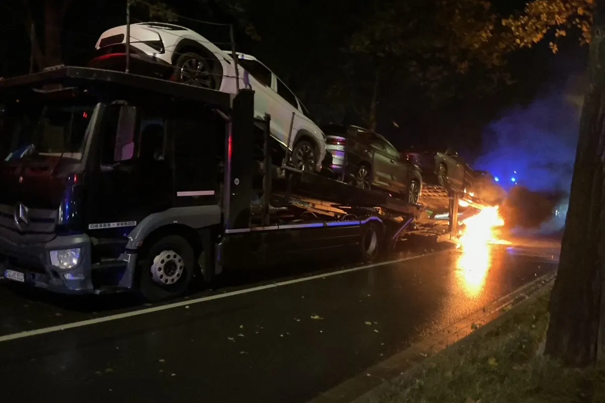 Ein Autotransporter mit mehreren Fahrzeugen darauf steht bei Dunkelheit auf einer nassen Straße. Ein Feuer brennt am Heck des Transporters.