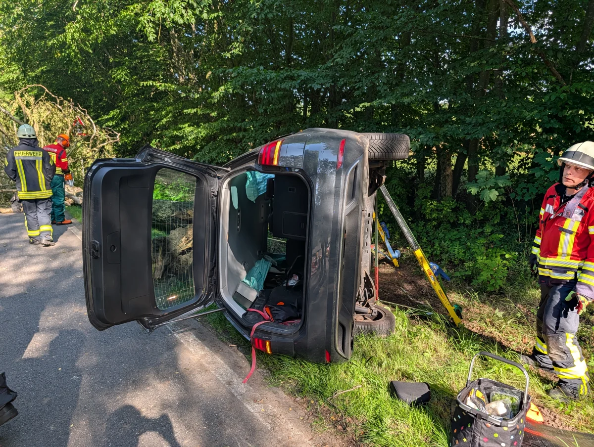 Ein Auto liegt auf der Seite am Straßenrand, während mehrere Feuerwehrleute in Einsatzkleidung die Unfallstelle sichern. Der Unfall ereignete sich neben einer Straße, die von Bäumen gesäumt ist.