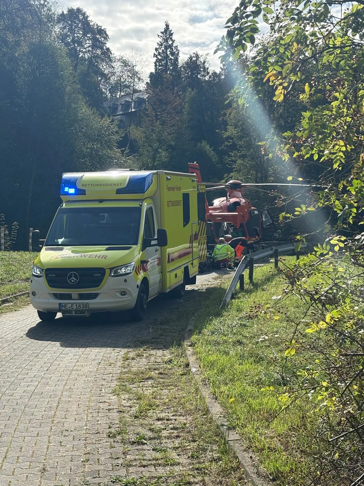 Ein gelber Rettungswagen steht auf einem gepflasterten Waldweg neben einem roten Rettungshubschrauber. Die Szene ist von Bäumen umgeben, und Sonnenlicht fällt durch die Blätter.