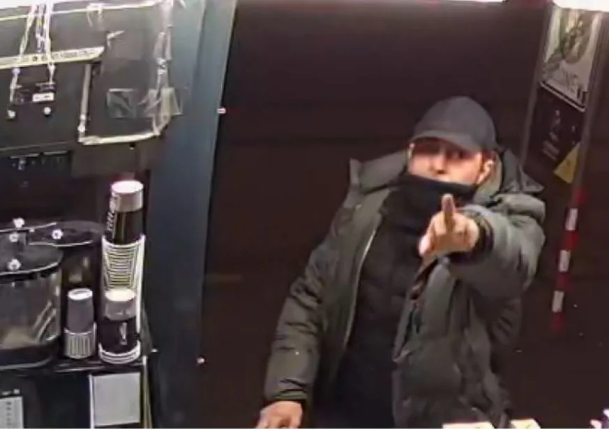 Ein Mann in dunkler Kleidung und Mütze zeigt auf eine Überwachungskamera in einem Raum, umgeben von Pappbechern.