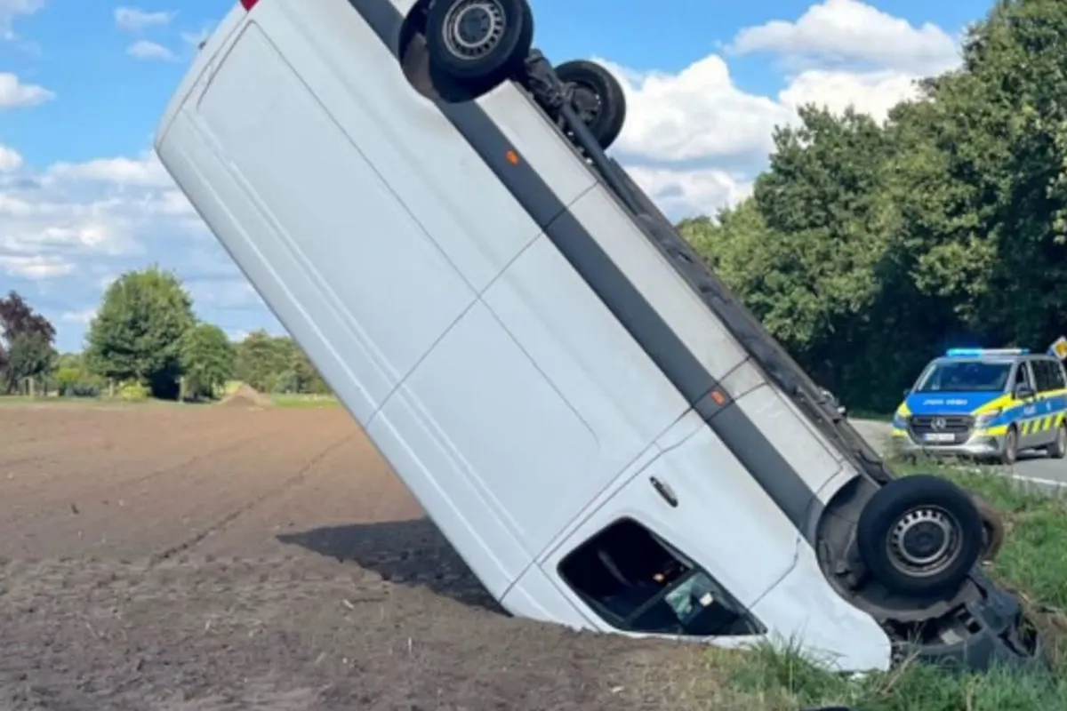 Ein weißer Lieferwagen ist auf einem Feld umgekippt, das Heck ragt in die Luft. Im Hintergrund steht ein Polizeifahrzeug an der Straße.