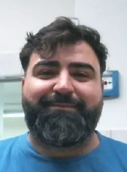 Ein Mann mit dunklem Haar und vollem Bart, der in einem Gebäude in die Kamera lächelt.