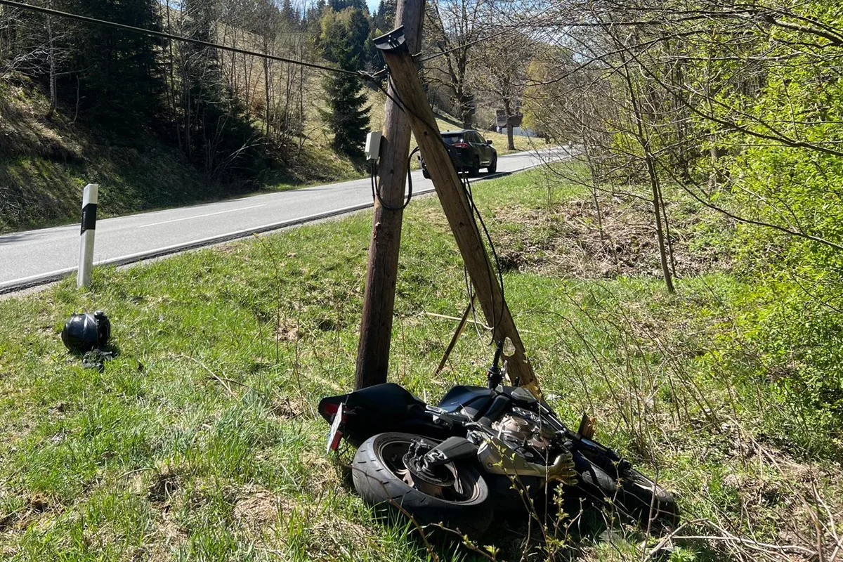 Ein Motorrad liegt neben einem Strommast auf einer Wiese. Im Hintergrund ist eine Straße mit einem Auto zu sehen.