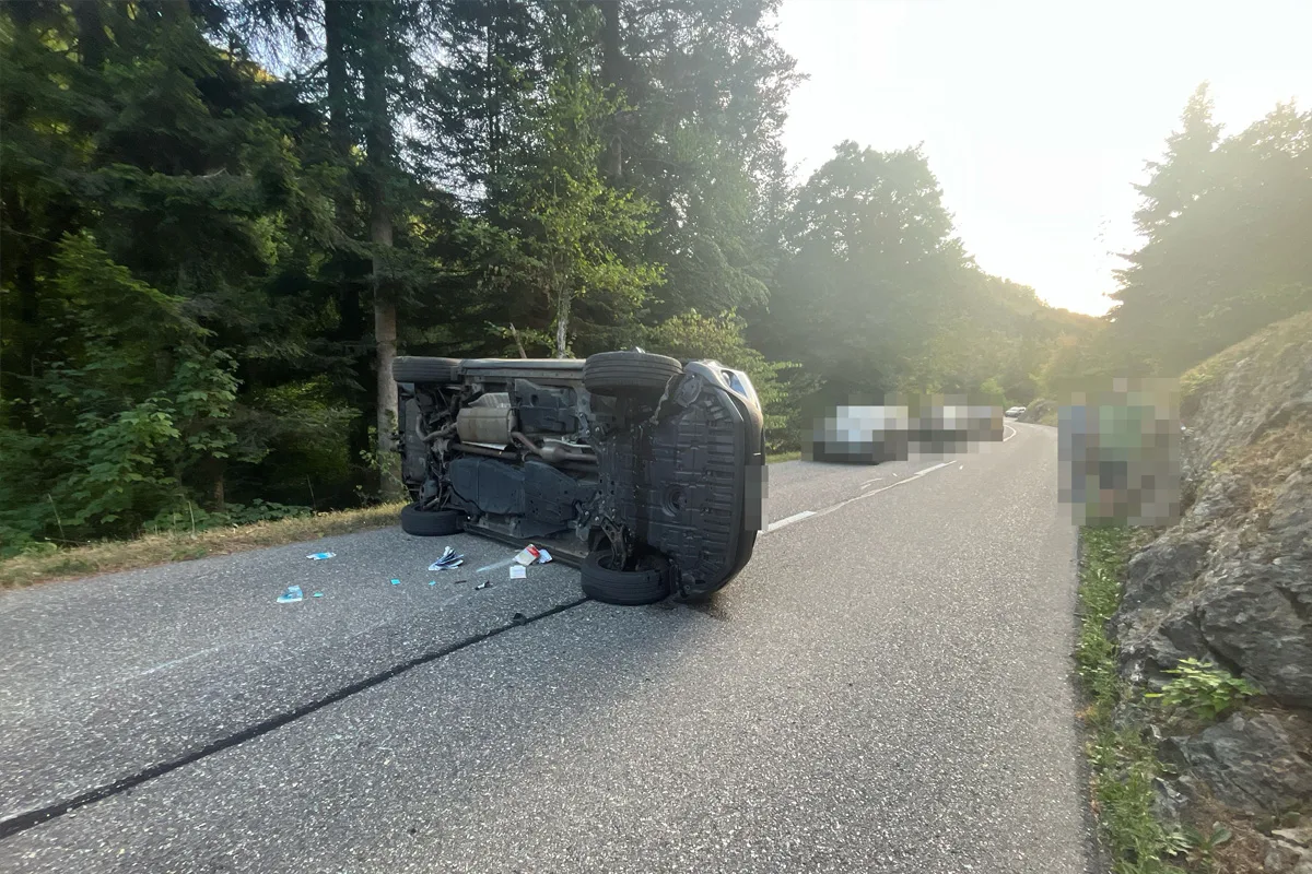 Ein Auto liegt seitlich auf einer asphaltierten Straße. Im Hintergrund sind Bäume und andere Fahrzeuge sichtbar.