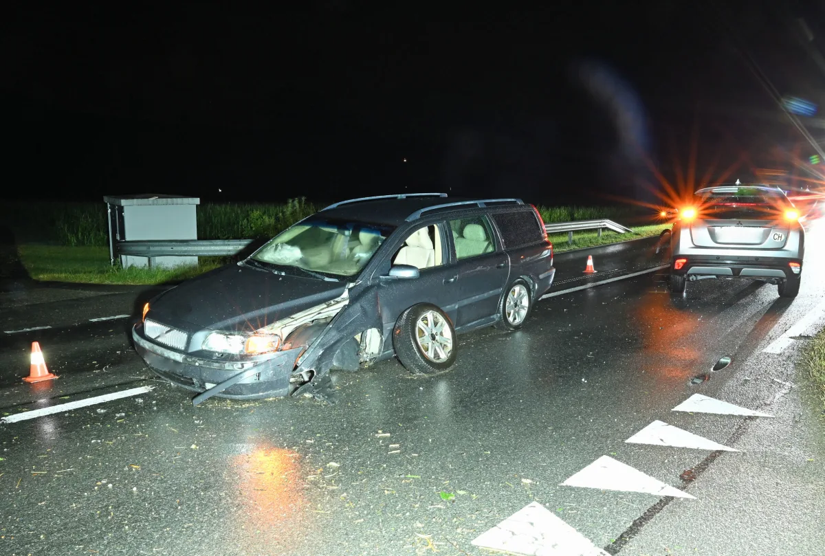 Ein stark beschädigtes Auto steht auf einer nassen Straße, daneben ein weiteres Fahrzeug. Es ist Nacht und es regnet leicht, mit Verkehrskegeln in der Umgebung.
