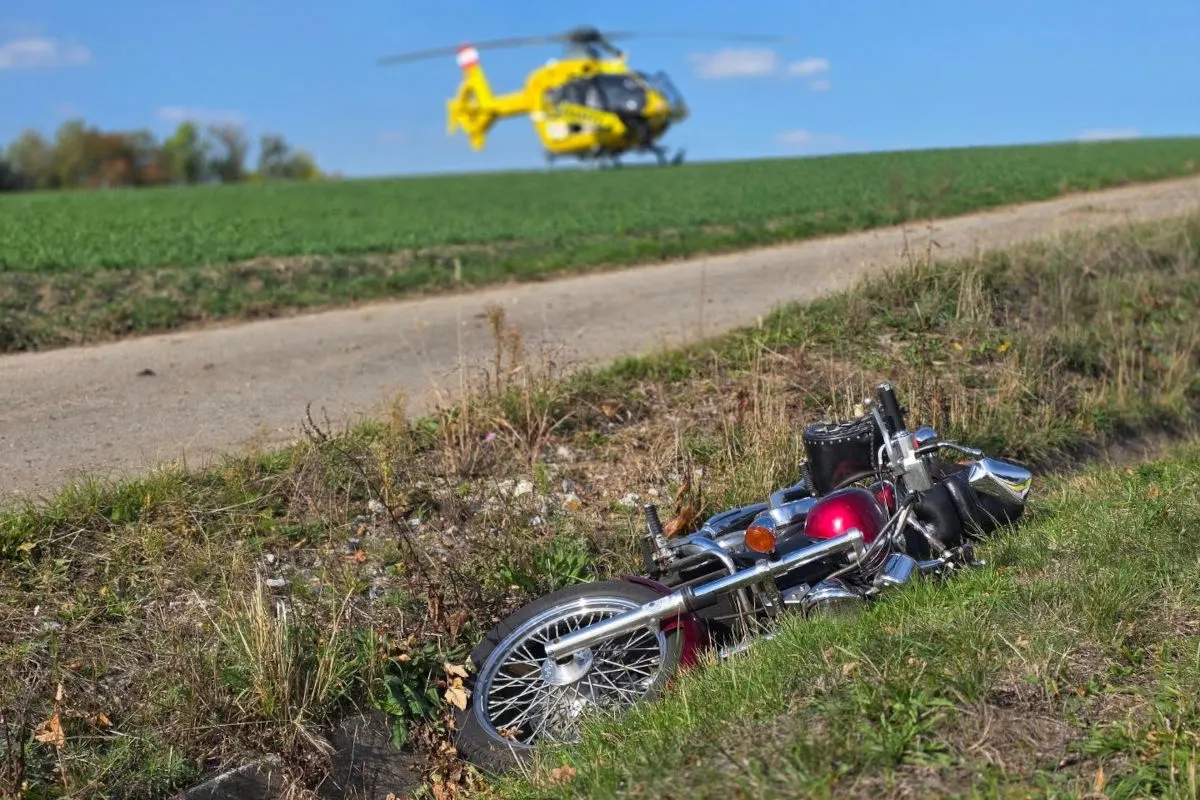 Ein rotes Motorrad liegt im Straßengraben, deutlich sichtbar im Vordergrund. Im Hintergrund wartet ein gelber Rettungshubschrauber auf einem Feld.