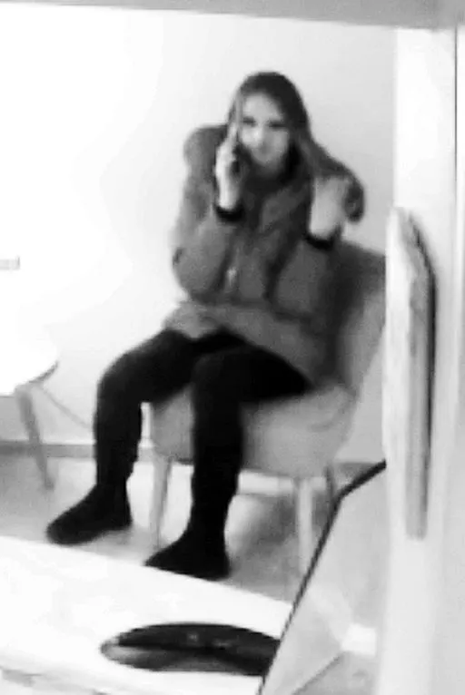 Eine Frau sitzt auf einem Stuhl und telefoniert. Das Bild scheint von einer Überwachungskamera aufgenommen zu sein.