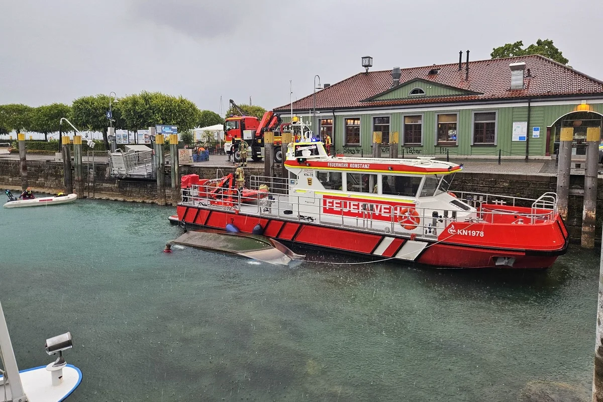 Ein auffälliges, rotes Feuerwehrboot mit der Aufschrift 'FEUERWEHR KONSTANZ' liegt im Hafen neben einem teilweise unter Wasser befindlichen Fahrzeug. Im Hintergrund sind Feuerwehrfahrzeuge und Gebäude sichtbar.