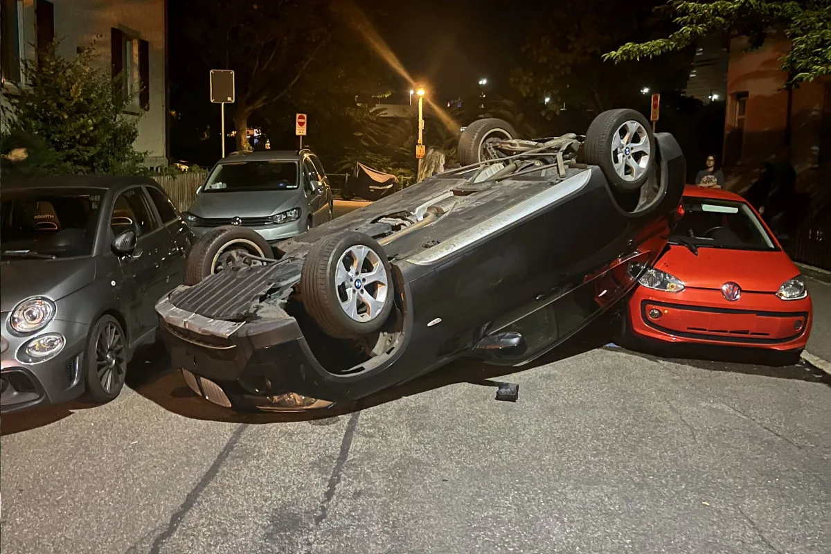 In einer Wohnstraße liegt ein dunkler Wagen auf dem Dach zwischen einem grauen Fiat und einem roten VW. Es ist Nacht.