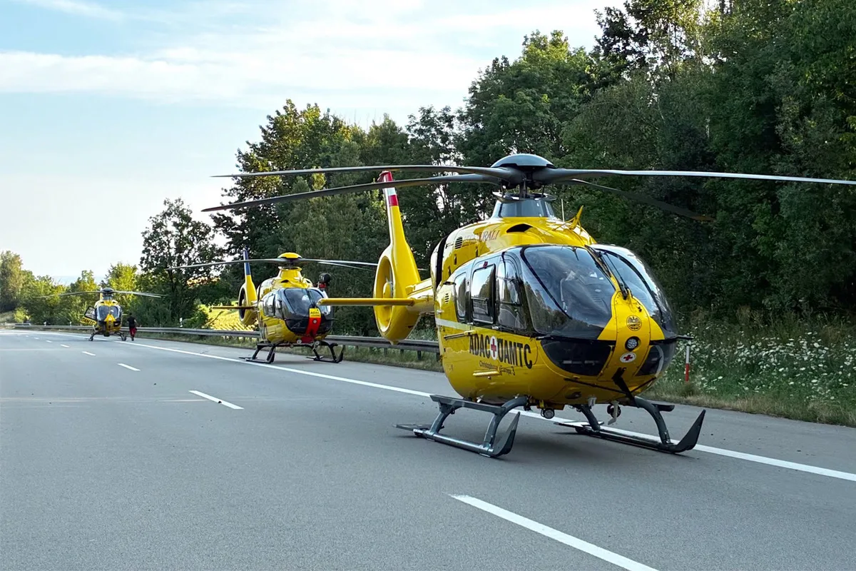 Auf einer Autobahn stehen drei gelbe Rettungshubschrauber in einer Reihe. Die Straße ist von Bäumen gesäumt, der Himmel ist teils bewölkt.