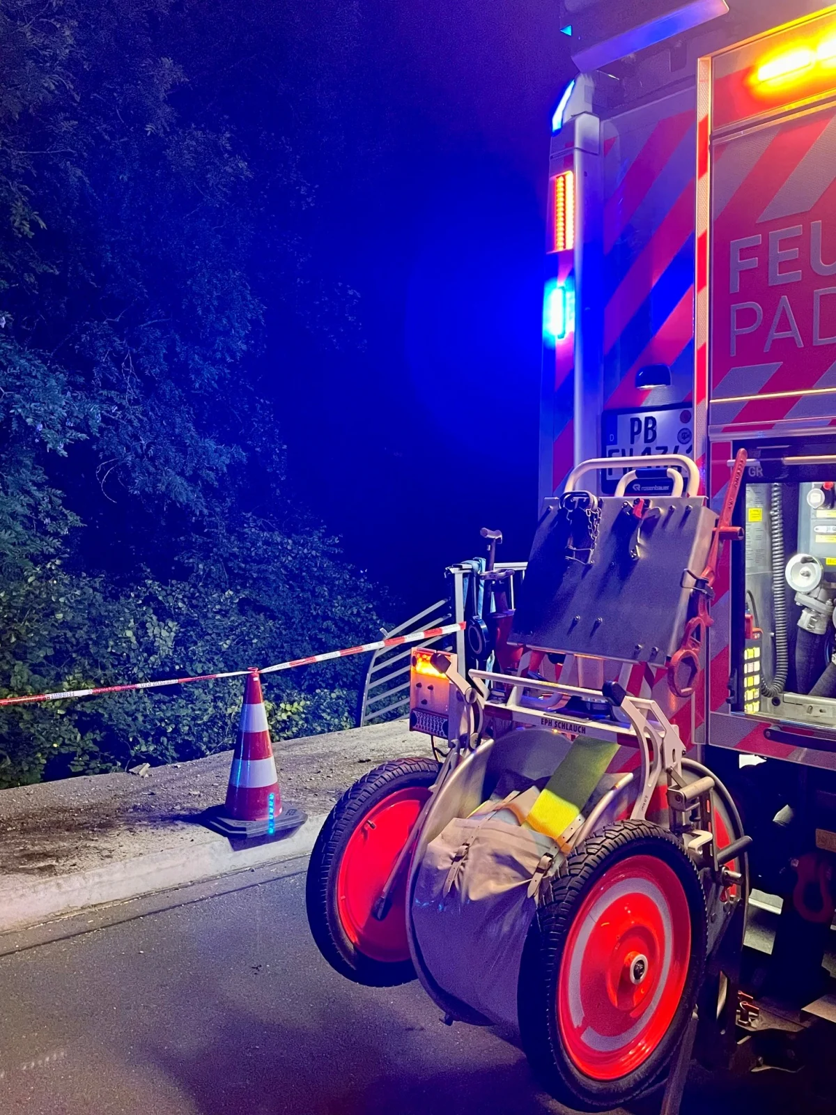 Ein Feuerwehrfahrzeug parkt nachts auf einer Straße, mit Absperrband und Pylon im Vordergrund. Ausrüstung ist am Heck des Fahrzeugs sichtbar.