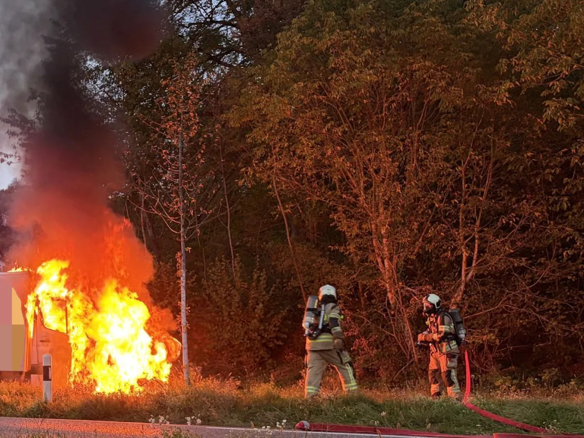 Zwei Feuerwehrleute in Schutzkleidung bekämpfen einen Fahrzeugbrand am Straßenrand bei dichten Rauchwolken.