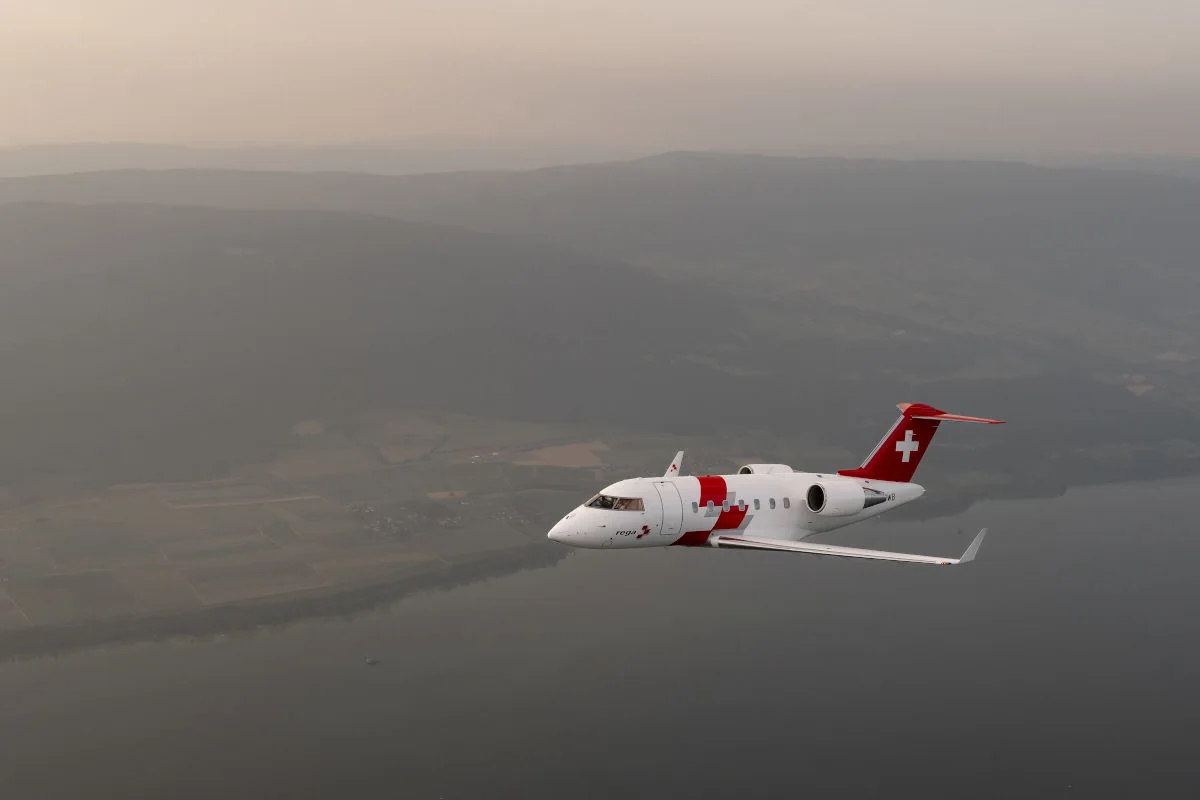 Ein rot-weißes Rega-Rettungsflugzeug fliegt über ein bewaldetes Gebiet mit Hügeln im Hintergrund und einem See im Vordergrund.