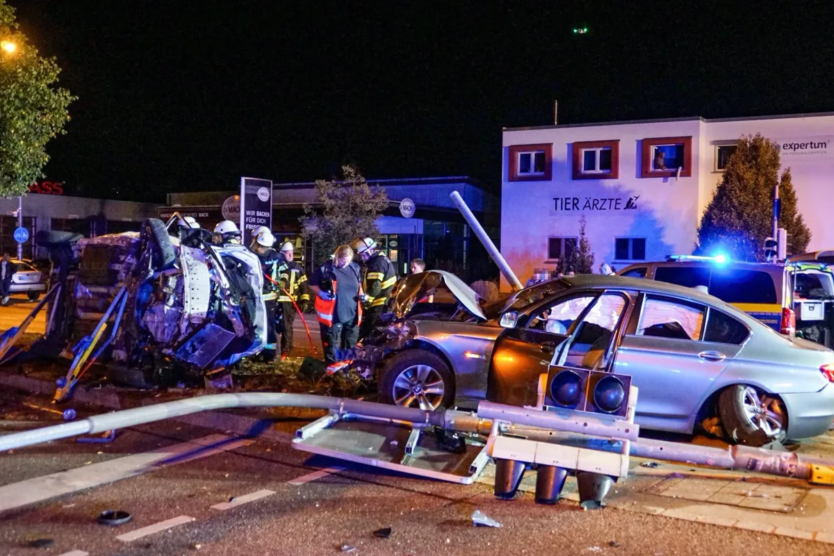 Zwei stark beschädigte Fahrzeuge nach einem Unfall, umgeben von Feuerwehrleuten bei Nacht. Eine Laterne liegt quer auf der Straße.